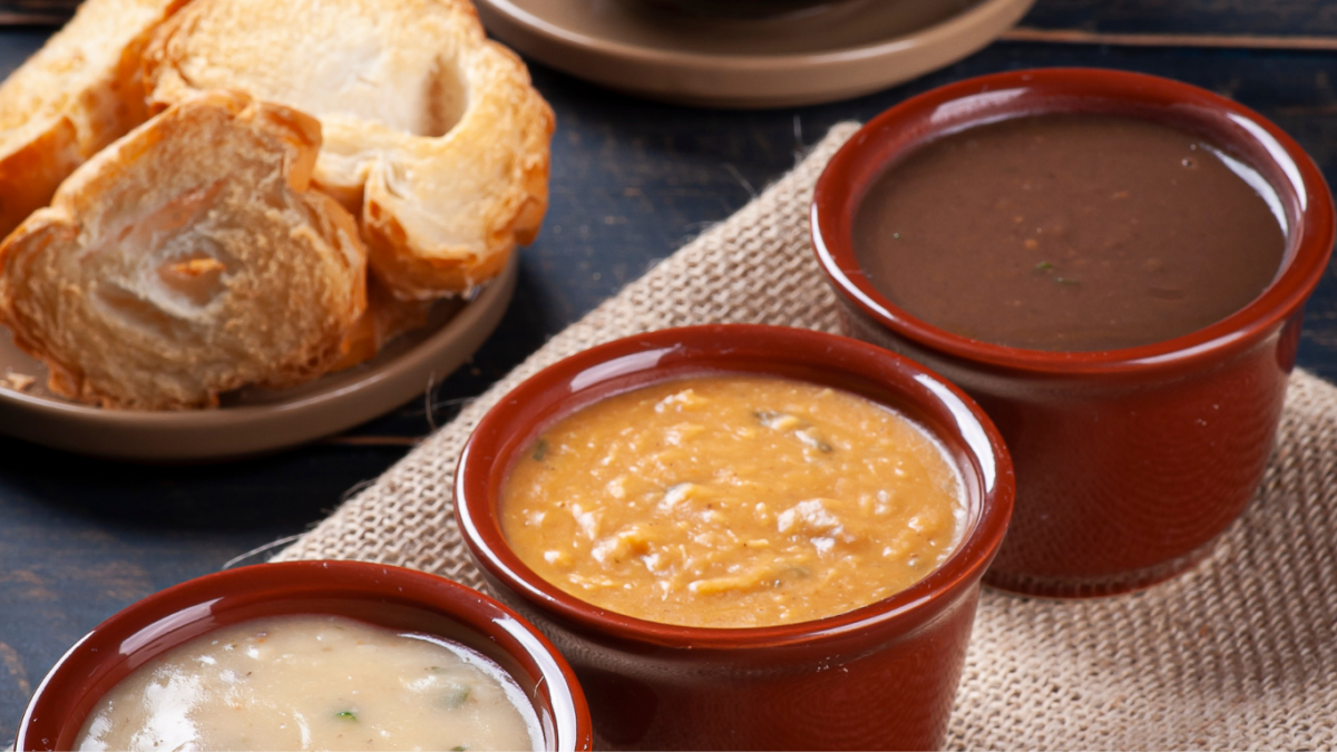 Mesa de festa junina com cumbucas de caldo fumegante, pão italiano de fundo sugerindo o consumo com as sopas
