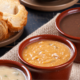 Mesa de festa junina com cumbucas de caldo fumegante, pão italiano de fundo sugerindo o consumo com as sopas