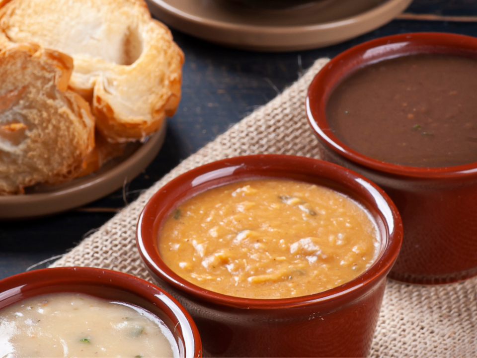 Mesa de festa junina com cumbucas de caldo fumegante, pão italiano de fundo sugerindo o consumo com as sopas