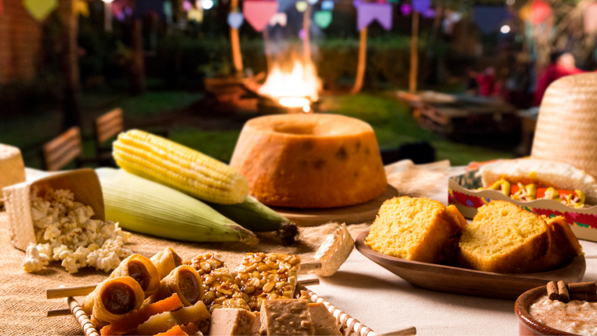 Mesa de festa junina com bolo fatiado, paçoca, pé de moleque, canjica e pipoca decorada com bandeirinhas coloridas e fogueira ao fundo